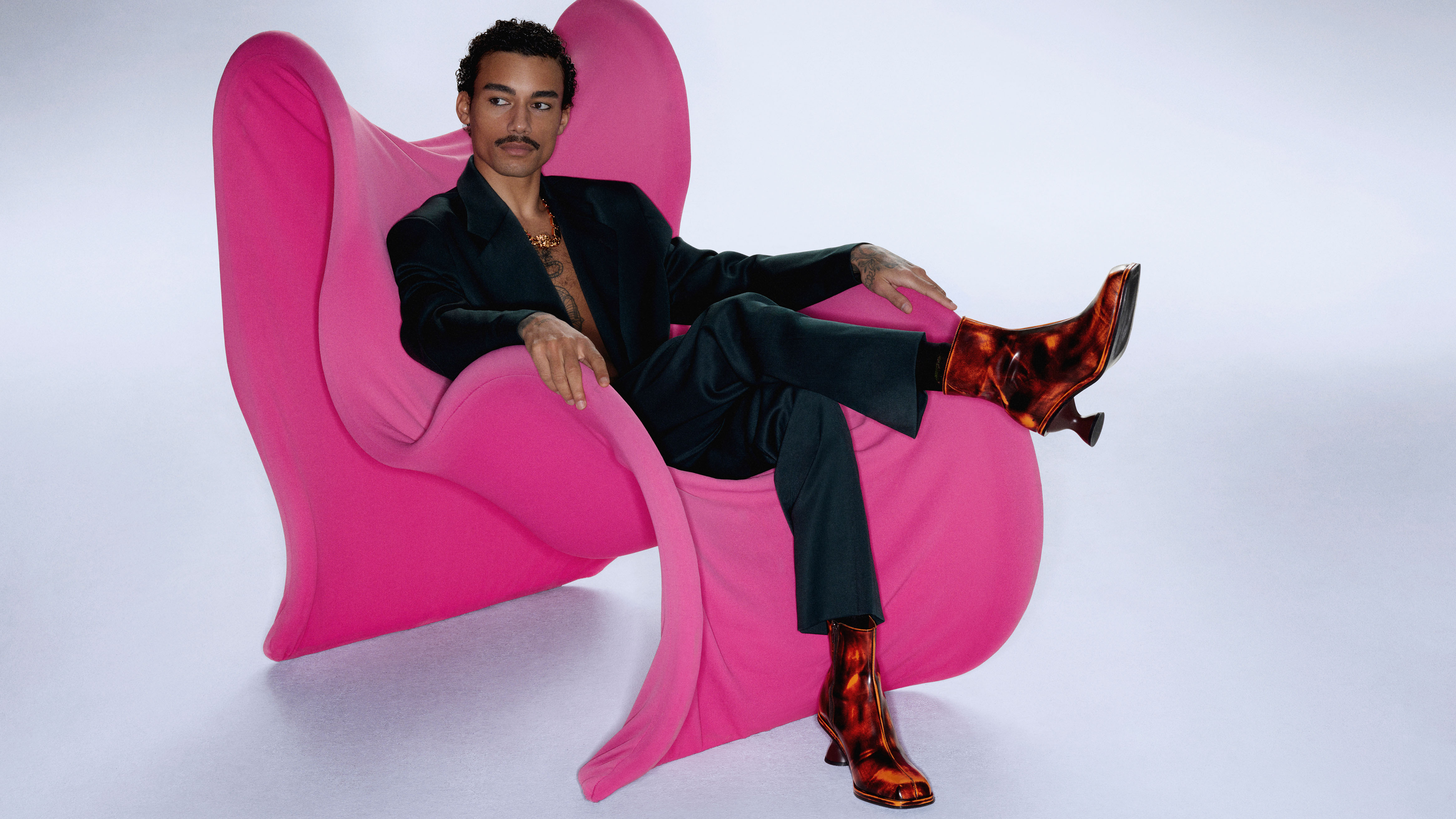 Fashion model sitting on a futuristic pink armchair wearing a black suit and characteristic boots