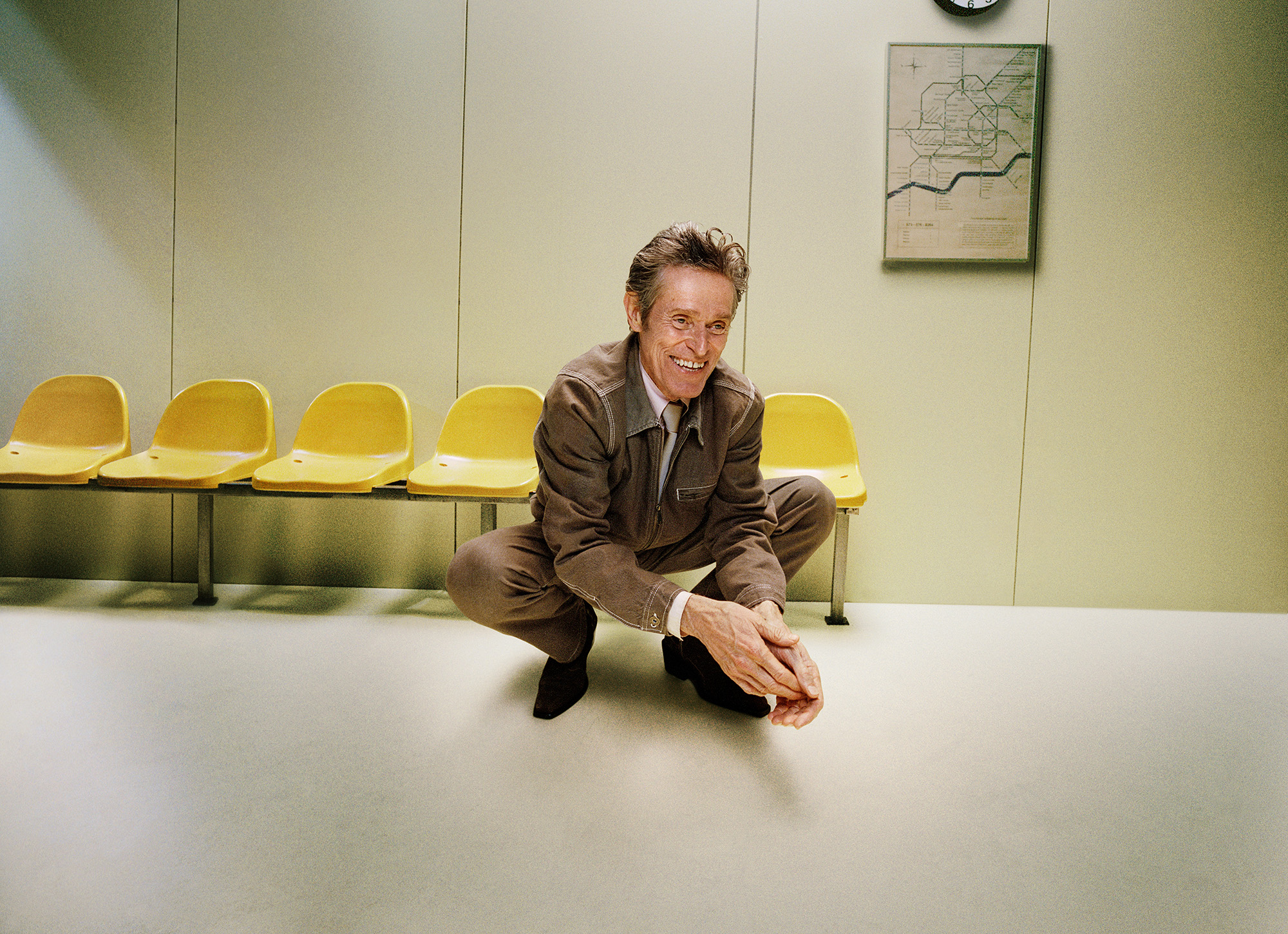 Willem Dafoe dressed in a brown suit crouching in front of a row of yellow chairs