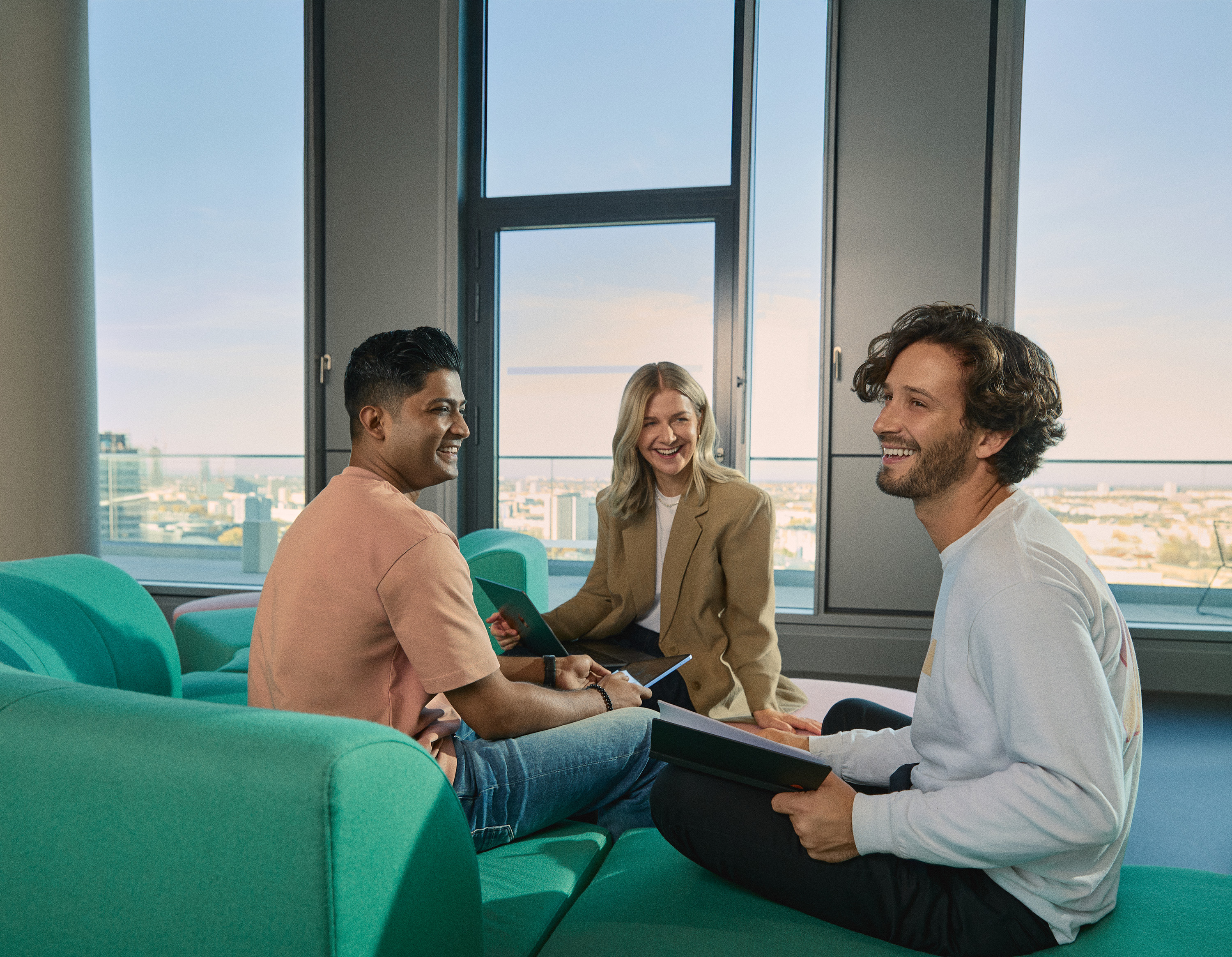 A group of three young professionals sits in a sunlit lounge area, engaged in a cheerful discussion. They are holding tablets and notebooks, suggesting a collaborative meeting. The background features large windows overlooking the city of Berlin, highlighting a modern and open workspace environment.