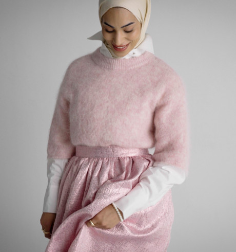 Das Model blickt lächelnd auf ihre Kleidung herab, sie trägt einen langen rosa Rock, einen übergroßen rosa Wollpullover und einen passenden beigen Hijab.