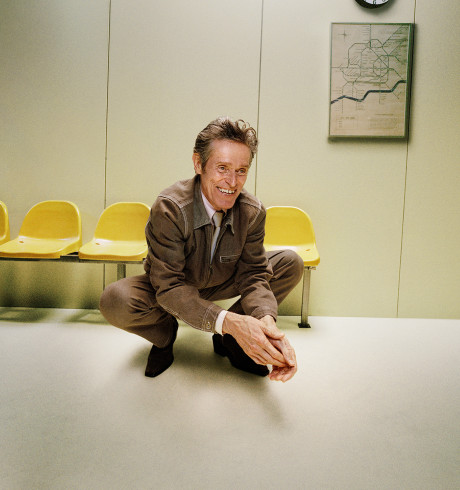 Willem Dafoe dressed in a brown suit crouching in front of a row of yellow chairs
