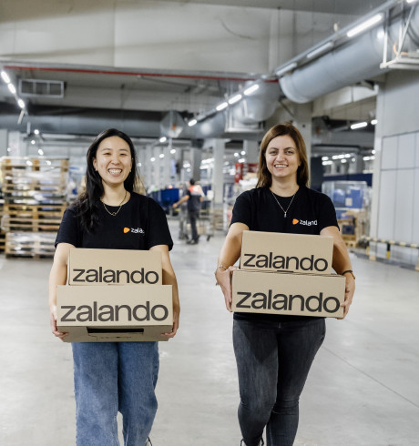 Vivian and Iva gehen mit jeweils zwei Zalando-Paketen durch eine Logistikhalle und lächeln in die Kamera