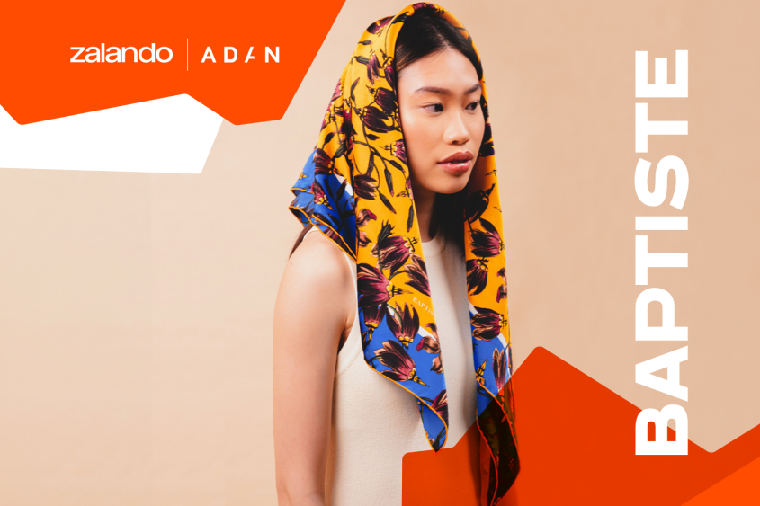 A woman is posing, wearing a simple white dress and a colourful headscarf, which is casually draped over the top of her head.