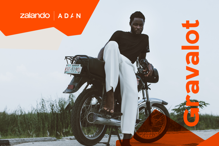 A man sits on the side of a motorbike, posing, wearing a black shirt and white trousers.