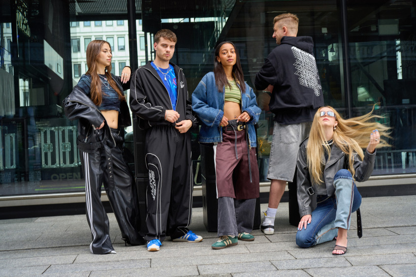A group of five young adults in cool, predominantly black streetwear posing on a street in front of a glass facade