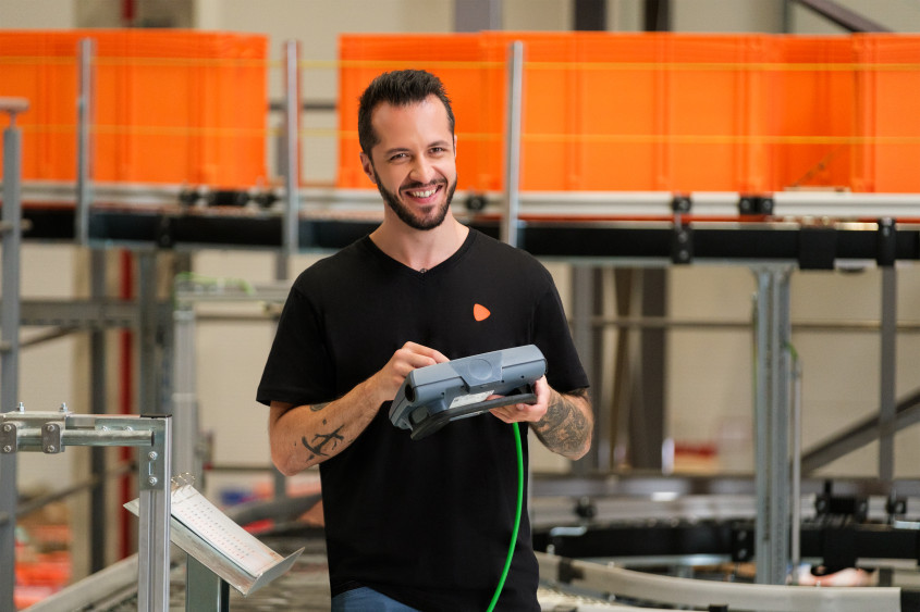 A smiling man in a black t-shirt stands in a logistics center, holding a grey handheld scanner. Large orange storage bins are visible on a conveyor system in the background.