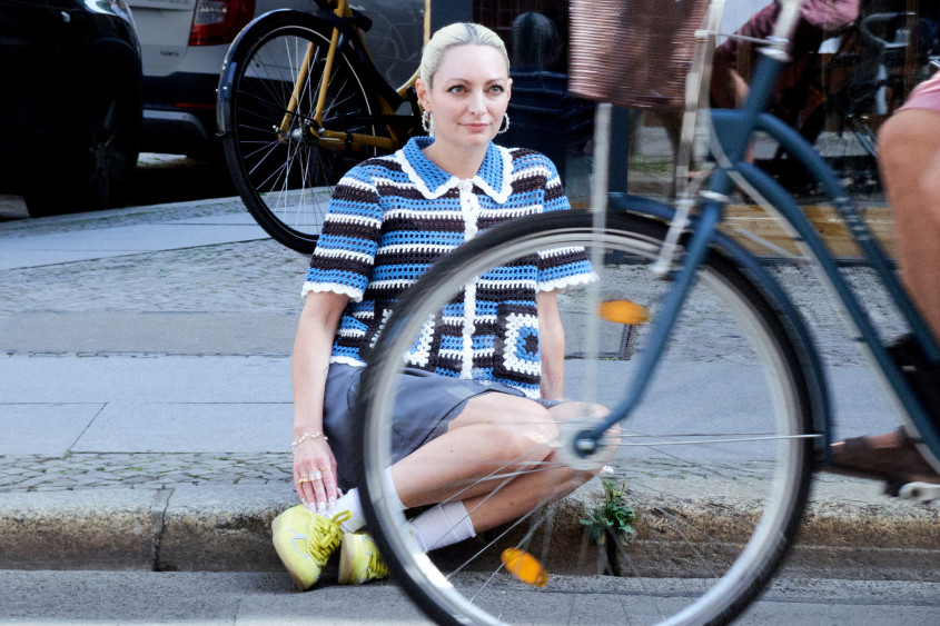 Young woman sitting on the curb while a bike is passing in front of her; she is wearing a blue white black striped crochet top, shorts and yellow sneakers
