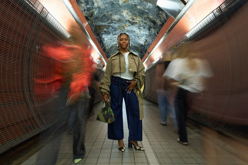 Black woman posing for the camera in what looks like a subway tunnel; she is wearing huge gold earrings, a short trenchcoat-style jacket, wide blue jeans and heels with a cow-hide pattern