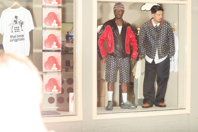 Two male fashion models standing in a shop window in cool streetwear; one man's shorts and the other man's jacket are showing the same specific pattern of light flowers on dark background