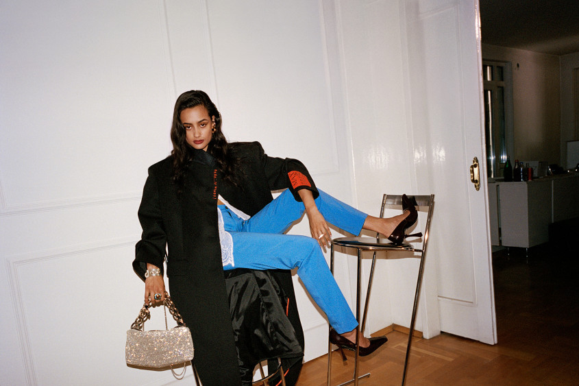 Female fashion model wearing bright blue trousers and an oversized black coat sitting casually on two barstools in what looks like an urban apartment