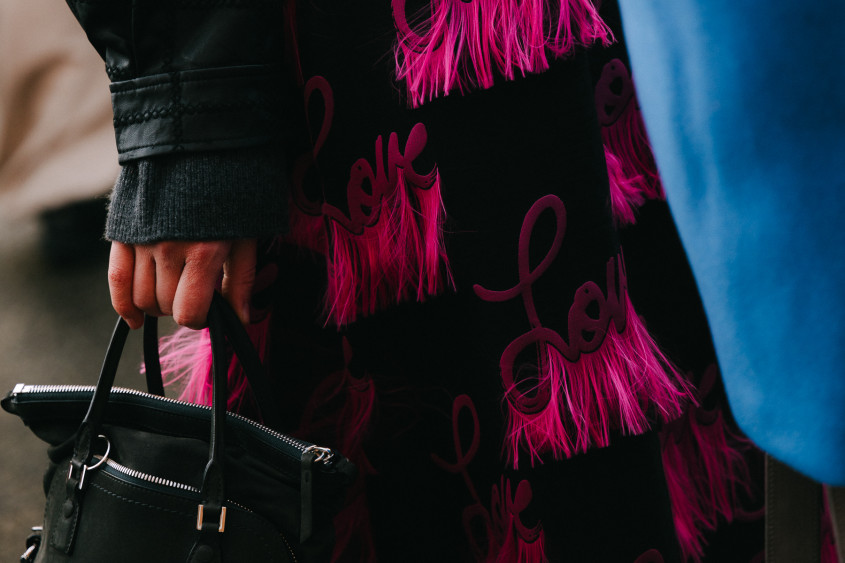 A person holding a black leather handbag; wearing a black coat with pink Love printed all over it