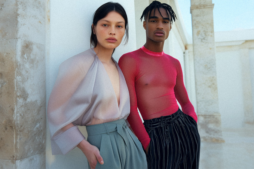 Two young fashion models posing outside in front of a white wall; their tops are of very thin material and slightly see-through