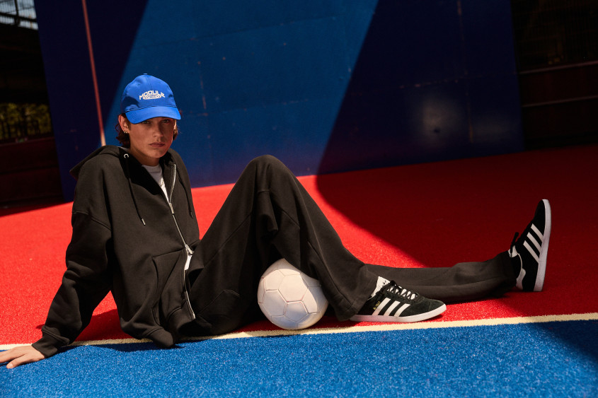 Fashion model wearing a blue basecap; black tracksuit, black and white Adidas sneakers; sitting on the ground of a blue and red sports area with a white ball