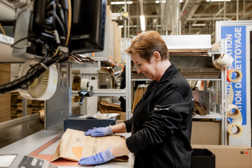 A side view of a smiling woman wearing blue safety gloves, carefully preparing a brown padded shipping envelope at a brightly lit warehouse workstation.