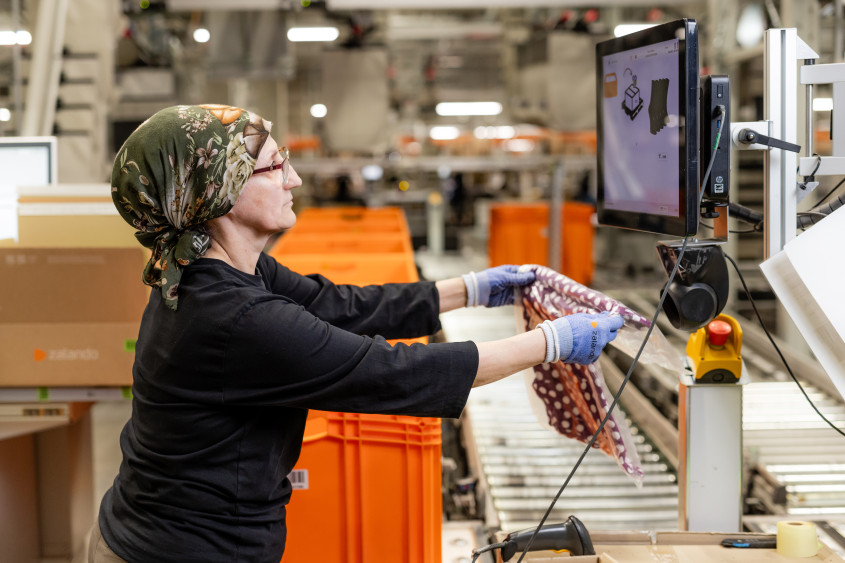 A woman wearing a green floral headscarf and glasses works at a packing station. She is holding a clear plastic bag containing a patterned garment while looking at a digital monitor displaying packing instructions.