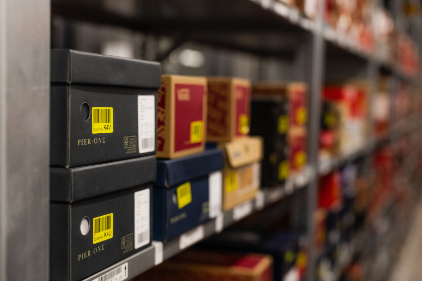 A shallow-focus shot of warehouse shelving filled with various shoeboxes. The boxes in the foreground are dark grey with PIER ONE branding and bright yellow barcode labels.