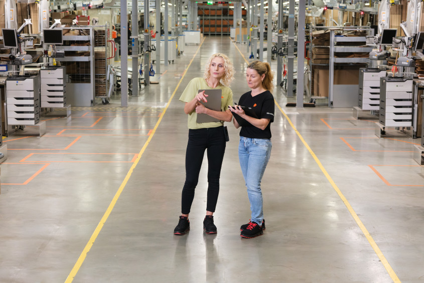 A wide shot of a large, modern warehouse aisle with two women standing in the center. They appear to be discussing something, holding a clipboard and a laptop, surrounded by automated packing stations.