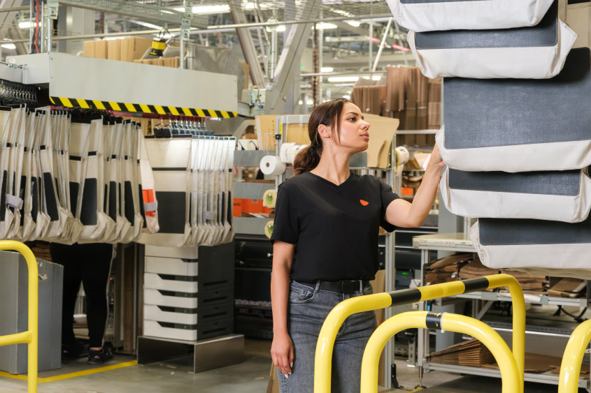 A woman in a black t-shirt looks upward while reaching for one of several white and grey shipping bags hanging from an automated overhead sorting system.