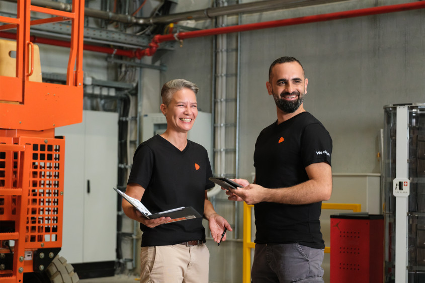 Two people in black Zalando t-shirts laugh together in an industrial setting. One person holds a clipboard while the other holds a small digital tablet. An orange industrial lift is visible in the background.