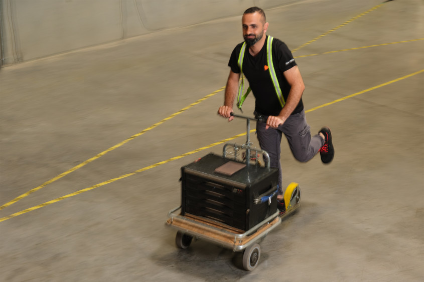 A man wearing a safety vest and a black t-shirt pushes a rolling cart carrying a large black tool chest across a warehouse floor marked with yellow lines.