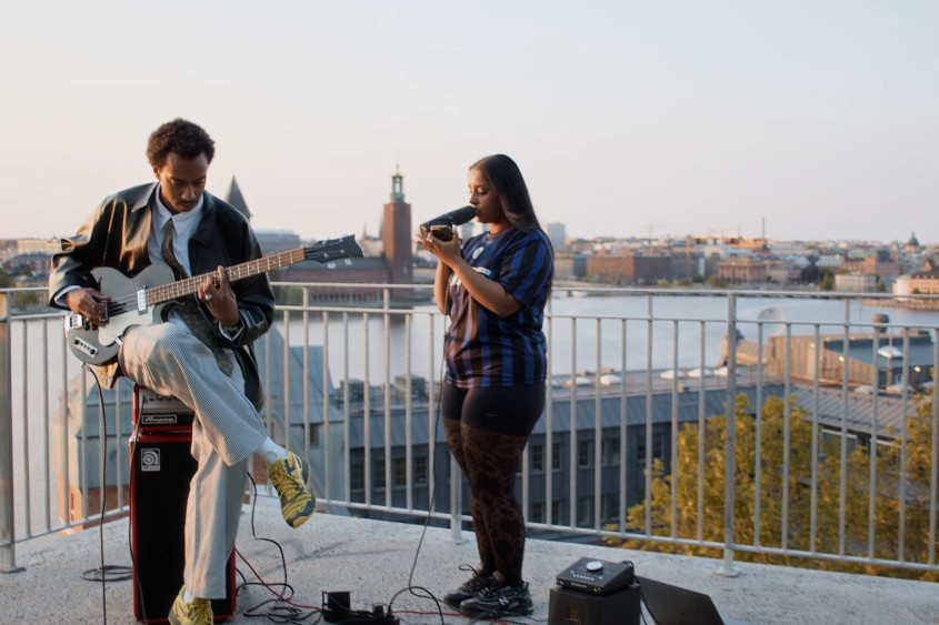 A woman sings and a man plays a guitar on a rooftop with a view of the city behind them
