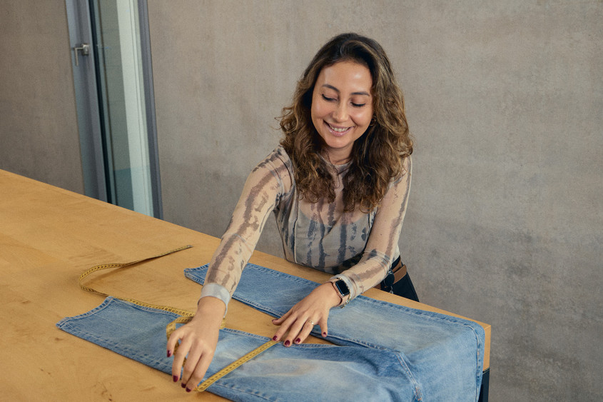 A woman with wavy hair, wearing a light patterned blouse, smiles as she measures blue jeans on a light wood workbench. She is holding a yellow measuring tape across the denim, working in a professional environment with a neutral gray wall.
