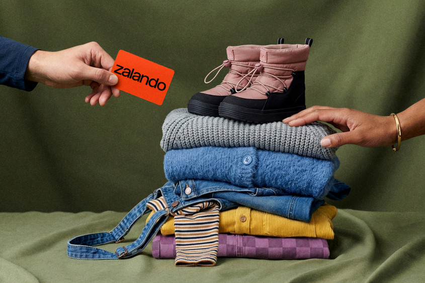 Ein Stapel bunter Kleidung; darauf ein Paar Kinderschuhe; von links kommt eine Hand mit einer Zalando-Karte