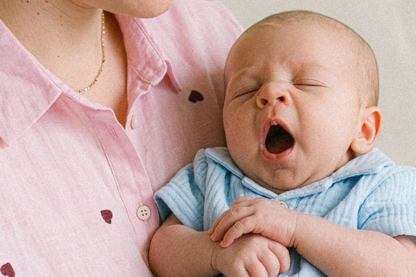 Yawning baby wearing a cute light blue musselin top on mother's arm