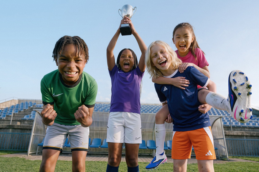 Four kids in sportswear in a small football stadium apparently celebrating a win, one is holding the cup in the air