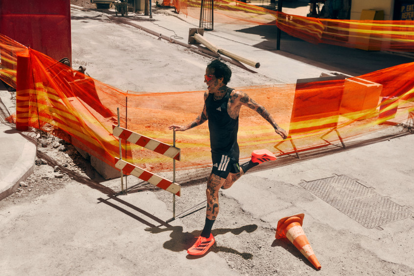 A tattoed runner with orange running shoes running along an urban construction site with orange barrier tape