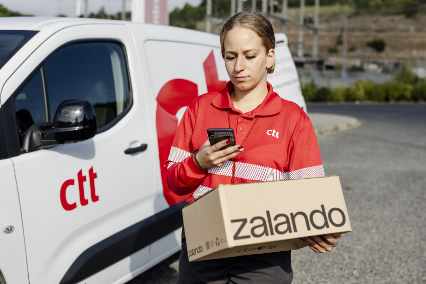 A ctt transport person with Zalando parcel in hand looking at her mobile phone next to her delivery van