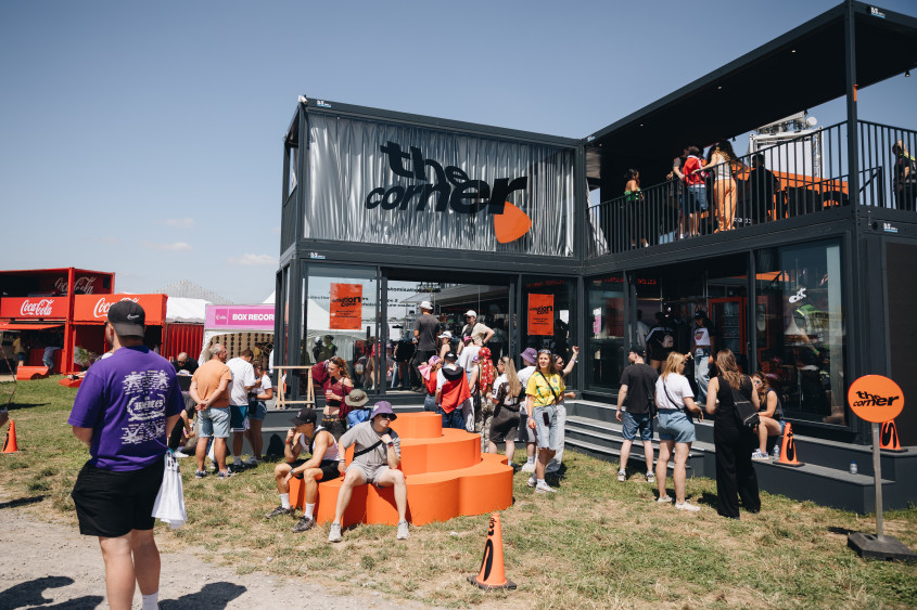 Festival-goers gather outside 'The Corner' pop-up in Belgium, a modern two-story black container building with orange block seating in front.