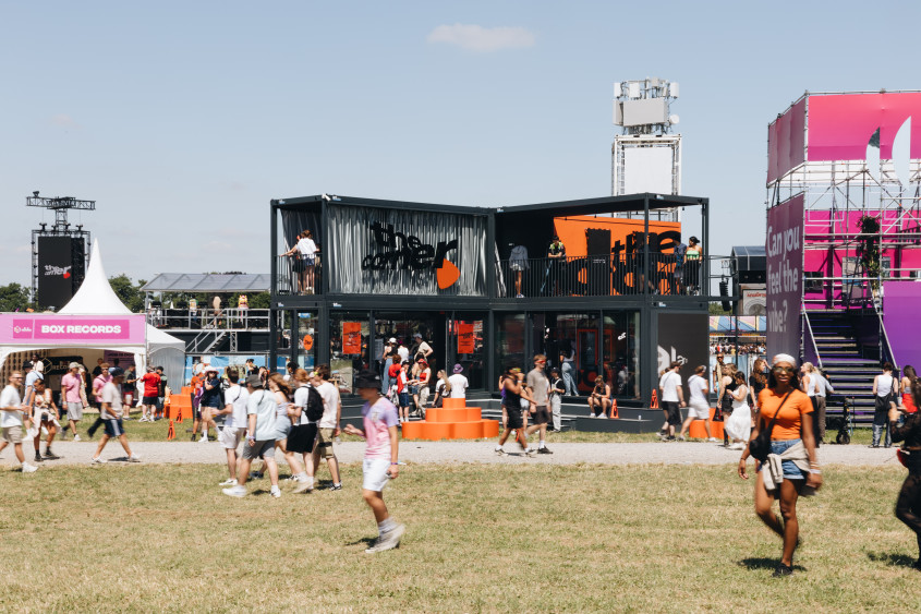 A wide view of 'The Corner' pop-up at a crowded music festival in Belgium, showing the two-story black building amidst a grassy field.