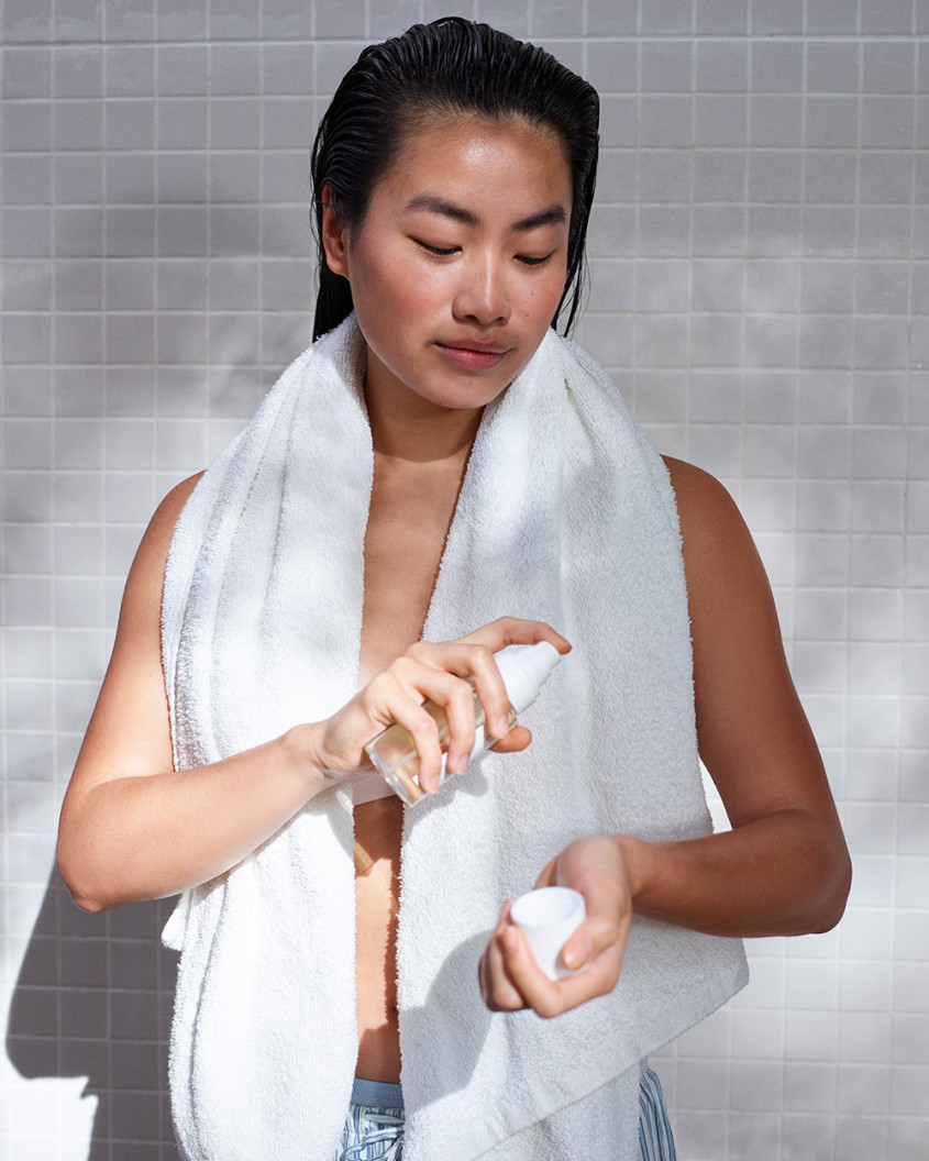 Person during beauty routine; long wet hair, white towel round their shoulders, spraying product onto wrist