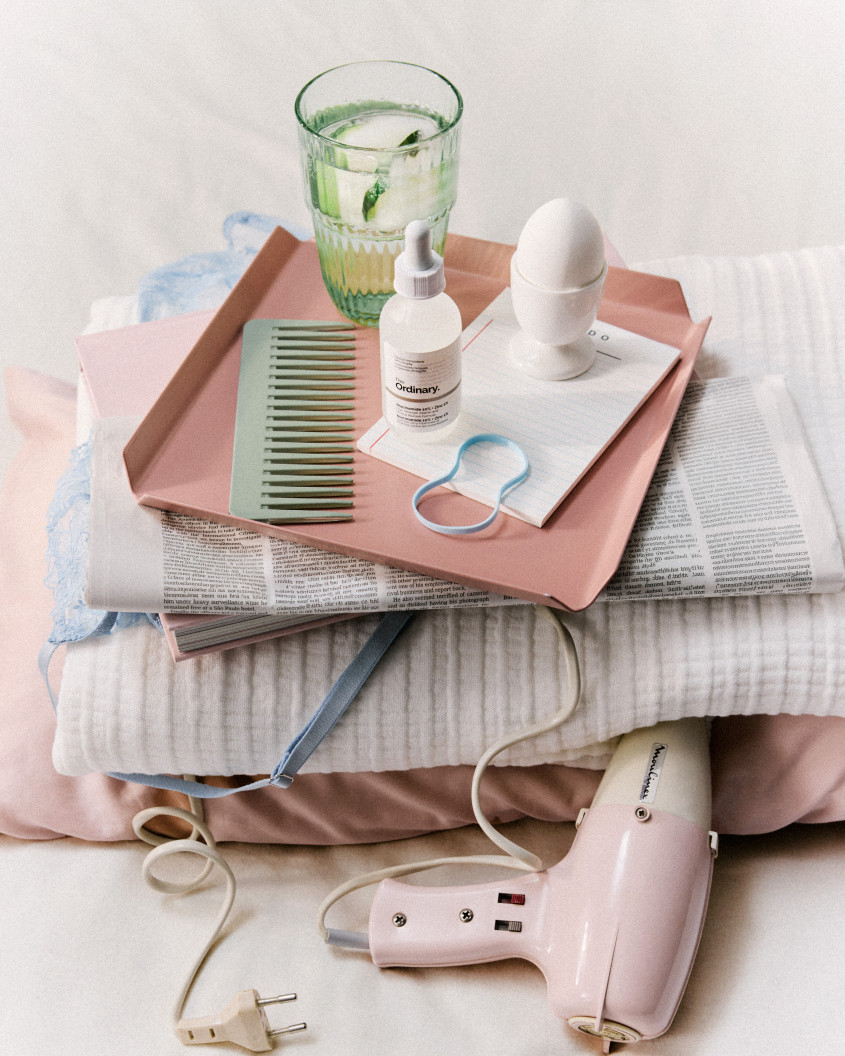 An aesthetically arranged breakfast tray with beauty products, a comb, an egg, a glass of water, resting on top of a newspaper