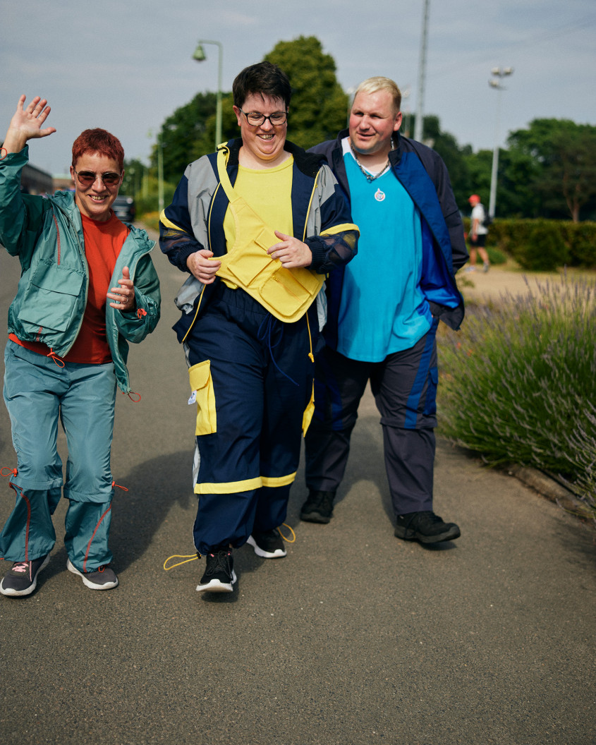 Three athletes in casual sports fashion walking on a path; one is waving to the camera