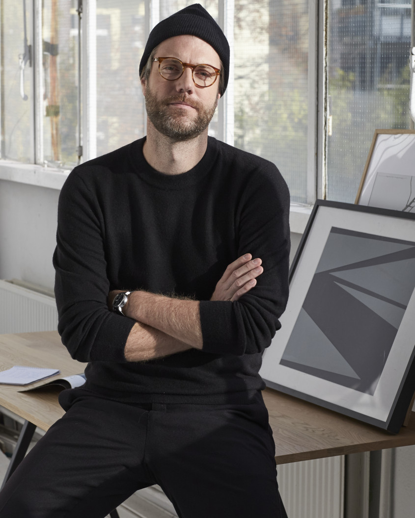 Portrait of Torsten Orendt in glasses and beanie sitting on a desk in front of a big glass window