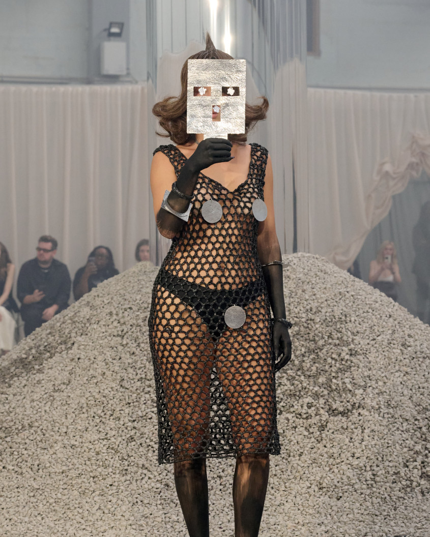 Fashion model in a net like dress and gloves, holding a metal mask in front of her face