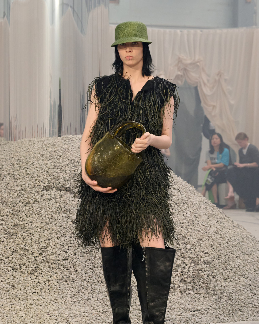 Fashion model in a furry dress, wearing a green hat, a bag made of glass and oversized boots