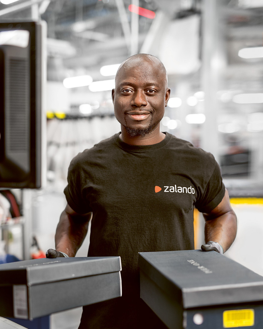 A Zalando employee in a warehouse setting holding two shoe boxes and smiling at the camera