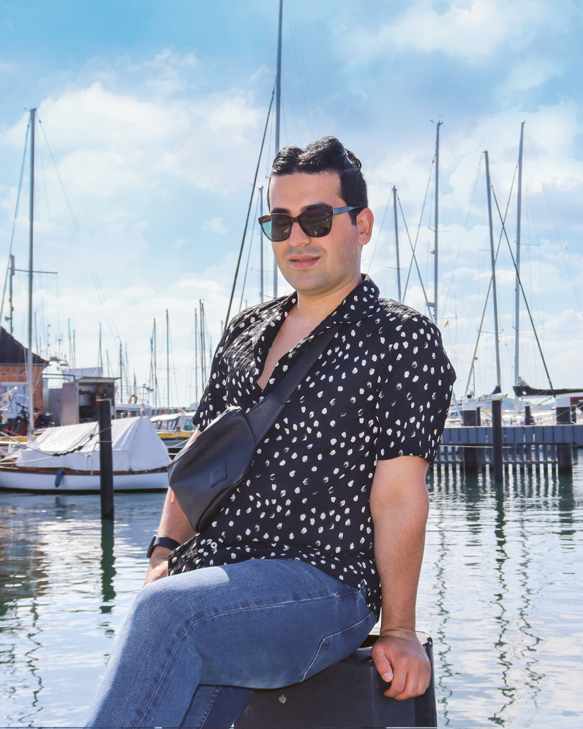 Man sitting at a harbour waterfront, wearing sunglasses on a sunny day; sailboats in the background