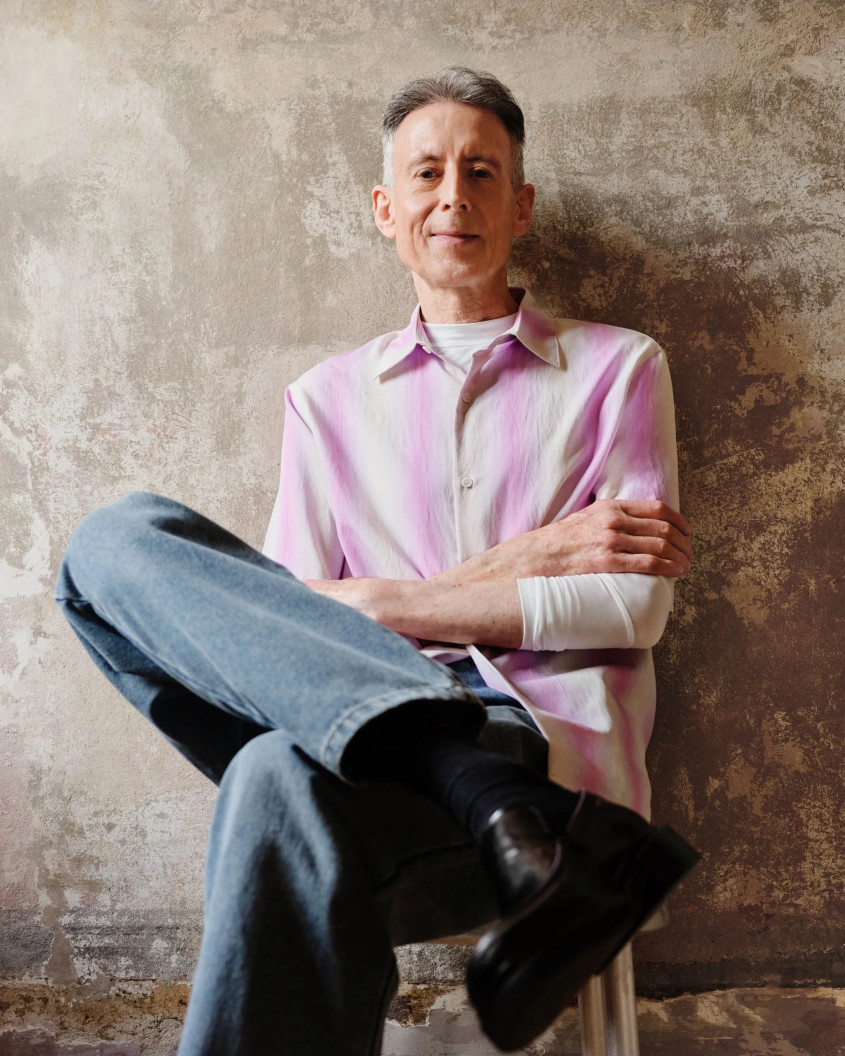 A grey man, sitting against a wall with one leg crossed, wearing a colourful striped button shirt and a blue jeans