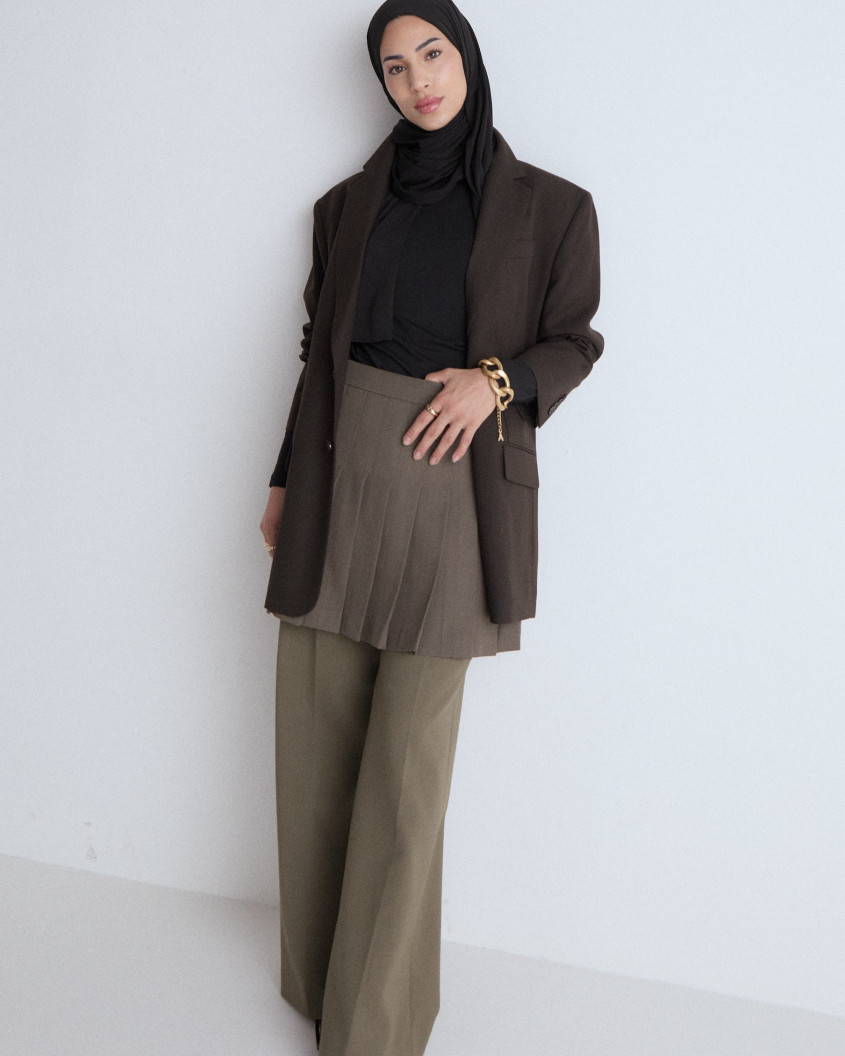 Full body picture of a model looking into the camera, wearing a short green pleated skirt over long trousers, a black top and oversized costume vest, combined with a golden statement bracelet and a black hijab.