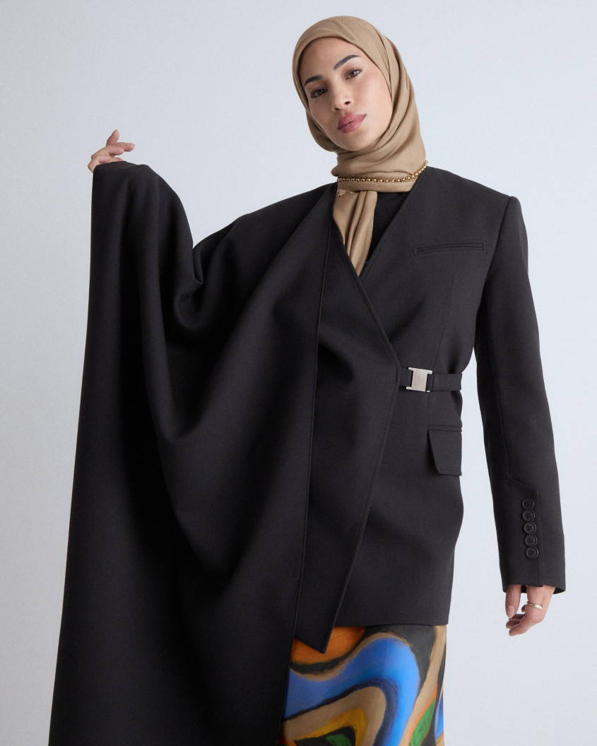 Model wearing a long colourful skirt, a brown hijab and a wide asymmetrical black cape, draped over her right arm.