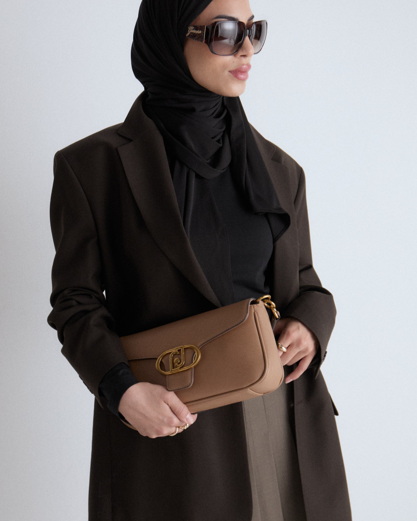 Close up of a model looking away, wearing a green pleated skirt, a black top and oversized costume vest, combined with a brown leather handbag, a black hijab and big sunglasses.