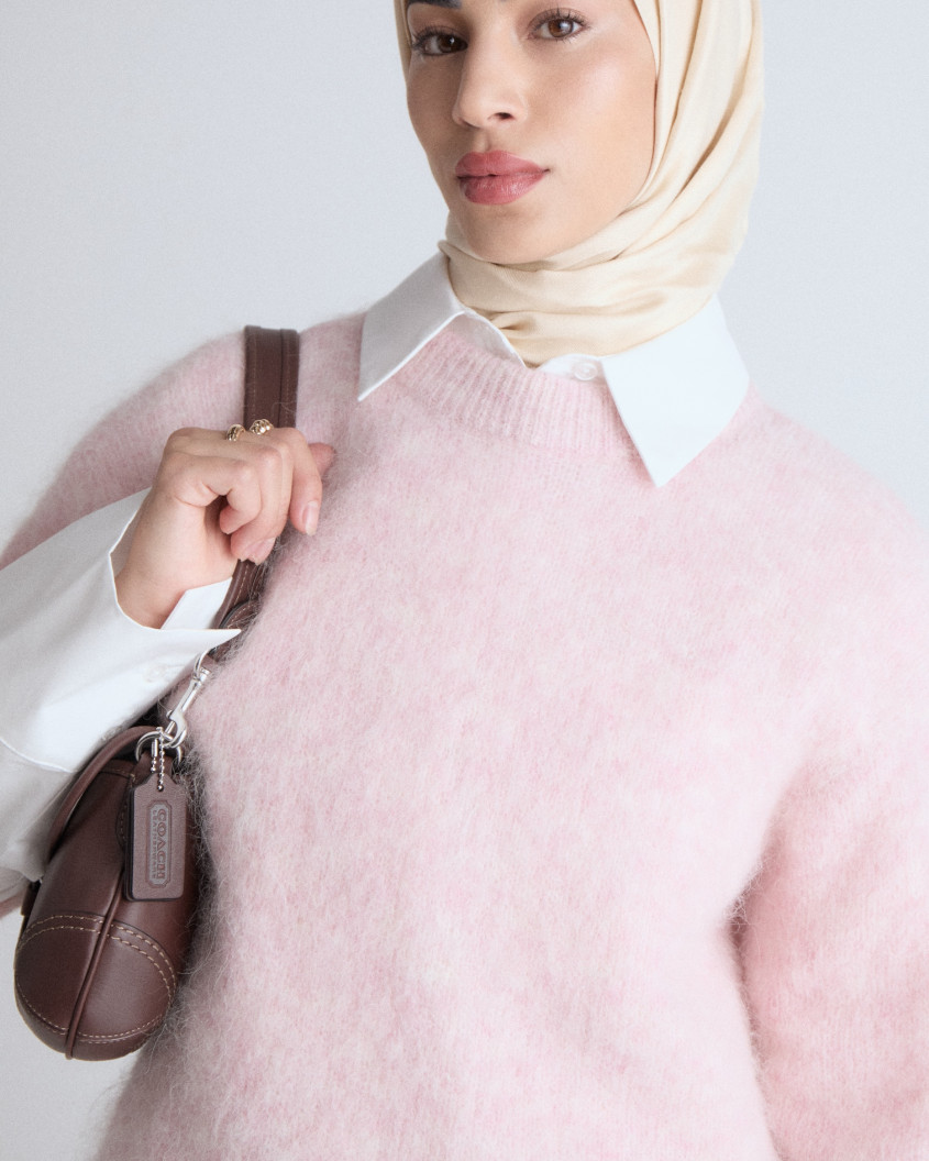 Close up of a model looking away, wearing a pink woolen oversized sweater over a formal white shirt, combined with a brown leather purse and matching hijab.