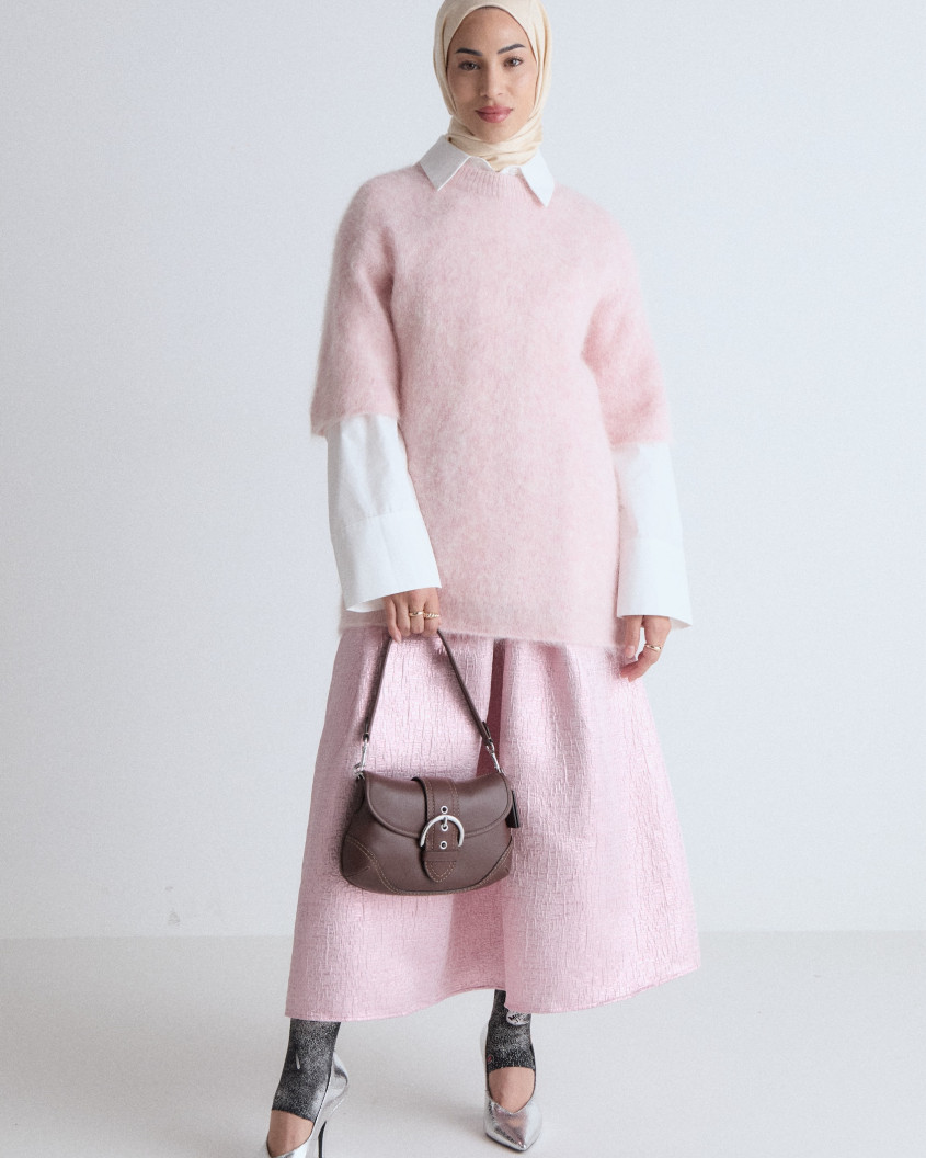 Full body picture of a model looking into the camera, wearing a pink long skirt, a pink woolen oversized sweater and matching hijab, combined with silver stiletto heels and brown leather purse.