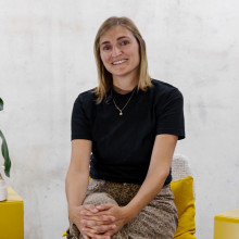 A woman with blond shoulder length hair is facing the camera sitting in a chair with her hands folded on her knee. She is wearing a black tshirt and grey trousers. She's surrounded by shoes from the Adaptive collection on yellow boxes and a huge plant.