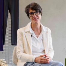 Woman with short black hair and glasses, sitting on a chair wearing a white shirt and creme blazer. She's surrounded by shoes and trousers from the Adaptive collection