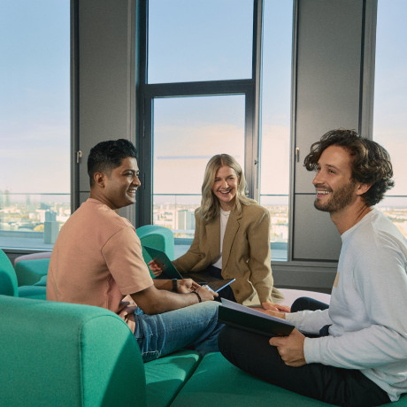 A group of three young professionals sits in a sunlit lounge area, engaged in a cheerful discussion. They are holding tablets and notebooks, suggesting a collaborative meeting. The background features large windows overlooking the city of Berlin, highlighting a modern and open workspace environment.